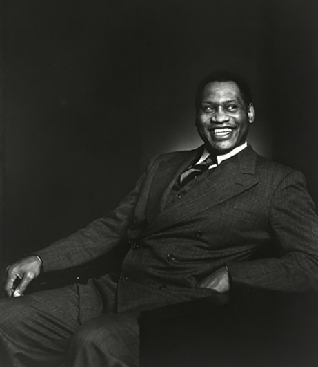 Iconic black and white photo portrait of a smiling Paul Robeson at roughly age 43, seated at an angle facing the camera, with both legs on the floor and his left arm on his chair’s armrest. He wears a dark suit jacket with wide lapels.