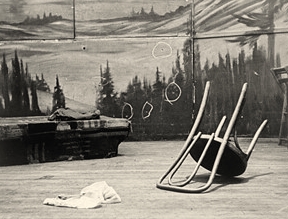 Black and white photo of the stage in the aftermath of Malcolm’s assassination. An upturned chair is in the foreground, the wall of the stage, with several bullet holes marked by chalked circles in the background.