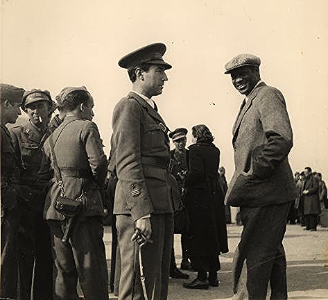 Black and white photo from 1938 showing, in the foreground, a smiling Paul Robeson, dressed in a suit, with his left hand in a pocket, wearing an English wool cap; and Robeson’s Spanish guide, Captain Fernando Castillo, dressed in a military uniform and hat, with his right hand on a walking stick.