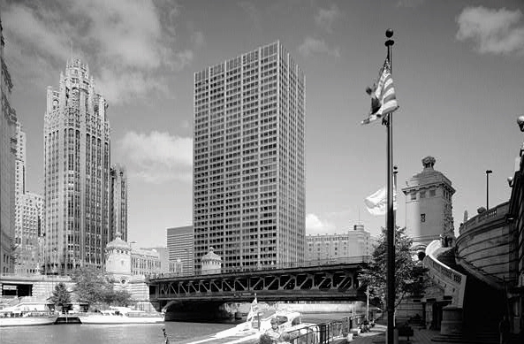 Black and white photo of tall buildings in Chicago. There is a flagpole flying an American flag, a river with some boats tied up along its edge. A staircase leads up to a steel bridge that crosses the river.