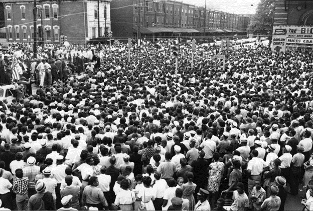 Black and white photo of a crowd of thousands gathered in the street facing a stage from which Dr. King addresses the multitude. A string of three-story rowhouses forms the backdrop of this image.