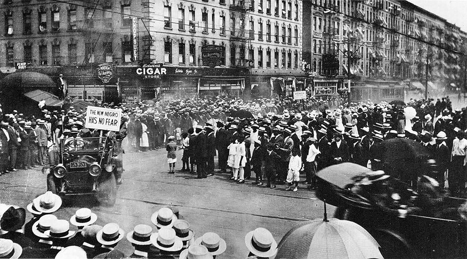 Black and white photo of the parade as it reaches an intersection in Harlem. Large crowds line the street. Parade participants ride in open cars in the foreground. A car shown turning at the intersection carries a plaque that reads THE NEW NEGRO HAS NO FEAR. A row of four-story tenement buildings, covered with fire escapes, form the background of the photo; a large white cigar sign appears on a tenement near the intersection.