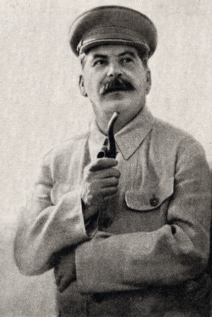 Iconic black and white photo of Joseph Stalin in 1937. He is dressed in a light-colored buttoned-down wool shirt, and he holds a pipe in his right hand at chest-level, with his left arm extended across his lower torso. He wears a military hat. The photo highlights Stalin’s heavy dark eyelashes and the thick mustache which covers his upper lip.