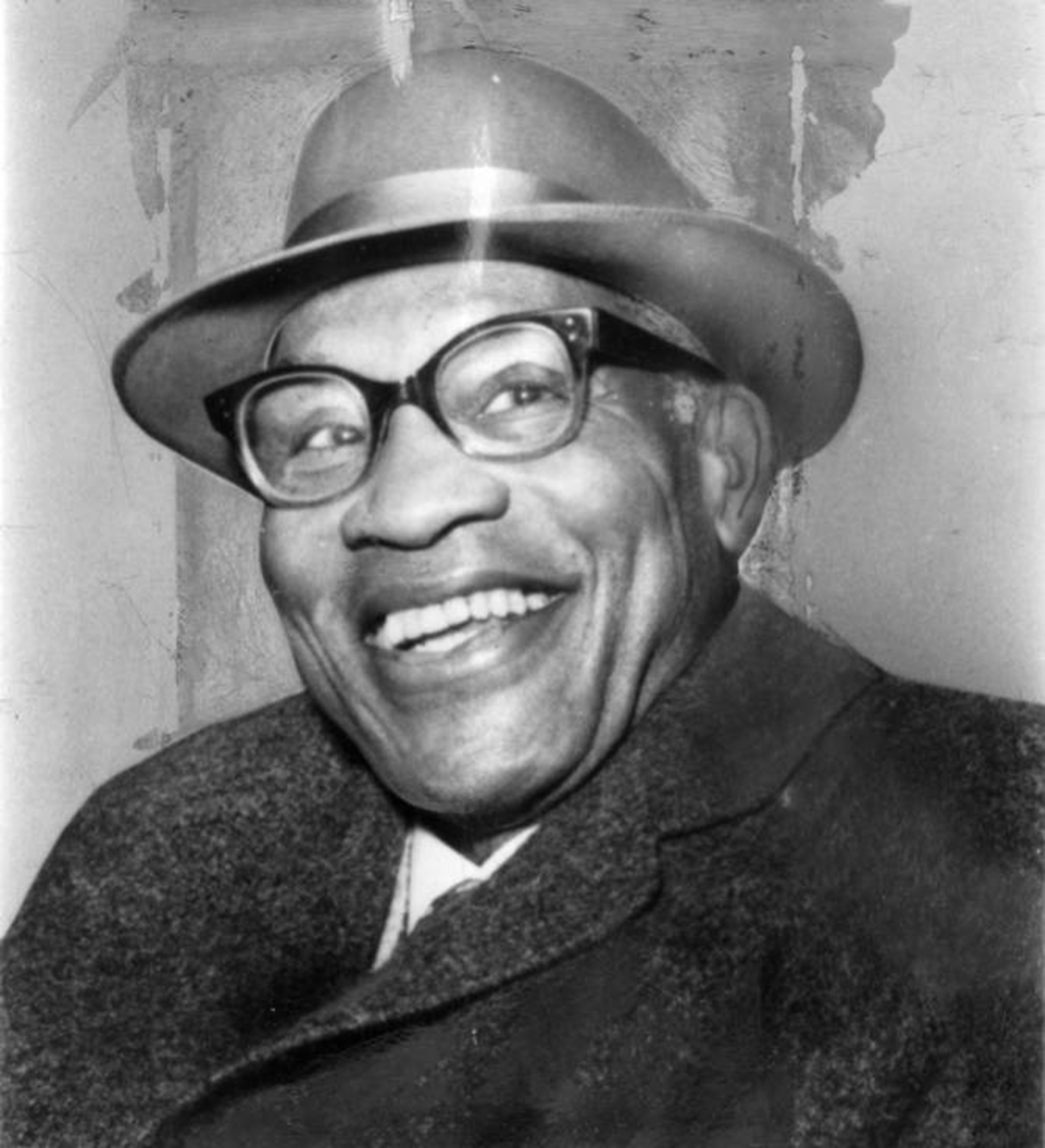 A candid black and white photo of a smiling Paul Robeson in his final years. Robeson’s pearly white teeth are prominent in the image, as are his large glasses. He is dressed in a dark wool winter coat and fedora.