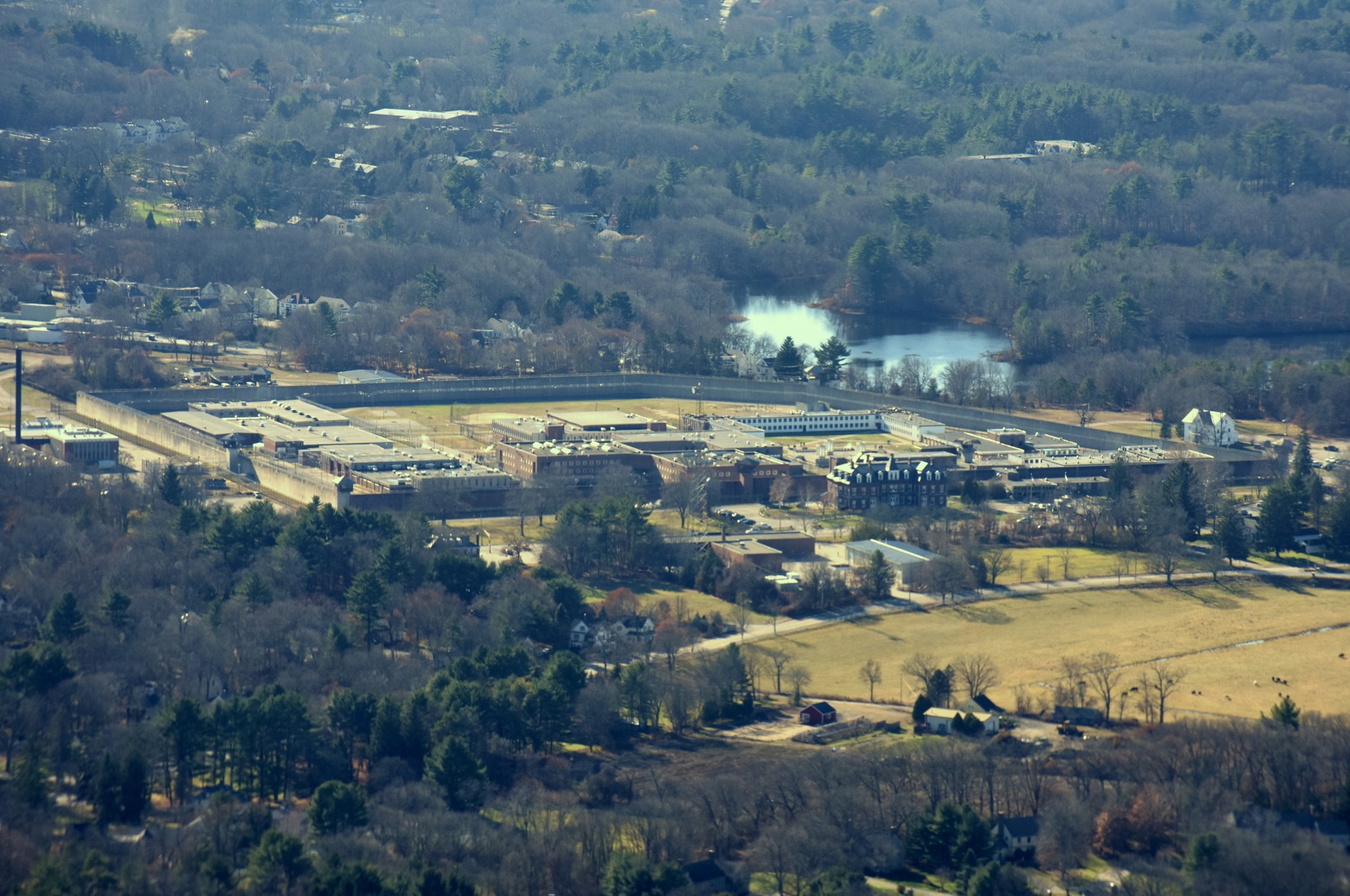 Color photo highlights the wall-enclosed prison facility, shown in the left-to-right center of the image, in its woodland location, with a river flowing just beyond the rear wall. The rest of the photo shows some unidentifiable low buildings enclosed by woods.