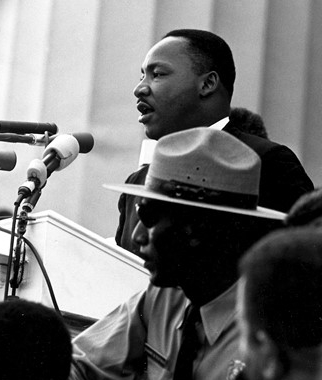 Black and white photo of Dr. Martin Luther King, Jr. speaking into a microphone with a state trooper near him