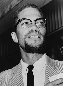 Black and white photo of Malcolm X in 1964. He is standing, wearing a light corduroy jacket, a white shirt, and a dark tie. His head is tilted to his left side. Emblematic of his break with the Nation of Islam, he now has a trimmed moustache connected to a small goatee, forming a circle.