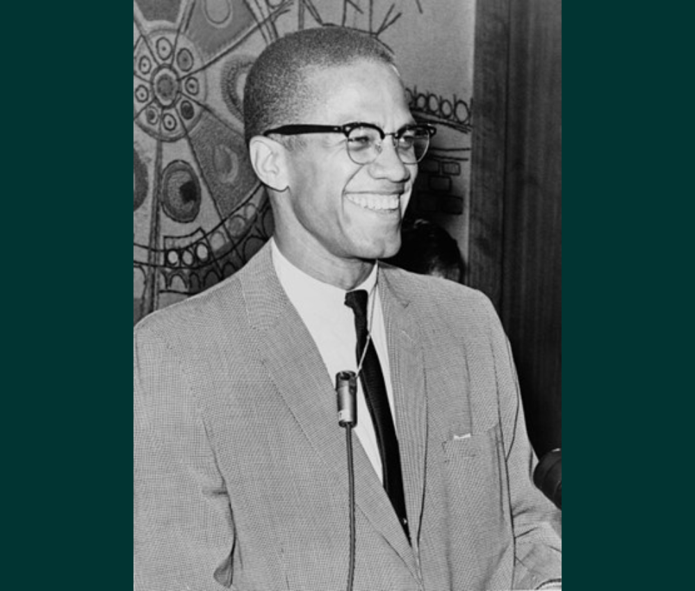 Black and white photo of Malcolm X in 1964. Shown smiling and relaxed, he stands at a microphone. He wears a light gray-tone suit or sports coat, with a dark tie. He has closely cropped hair, is clean shaven, and wears glasses.