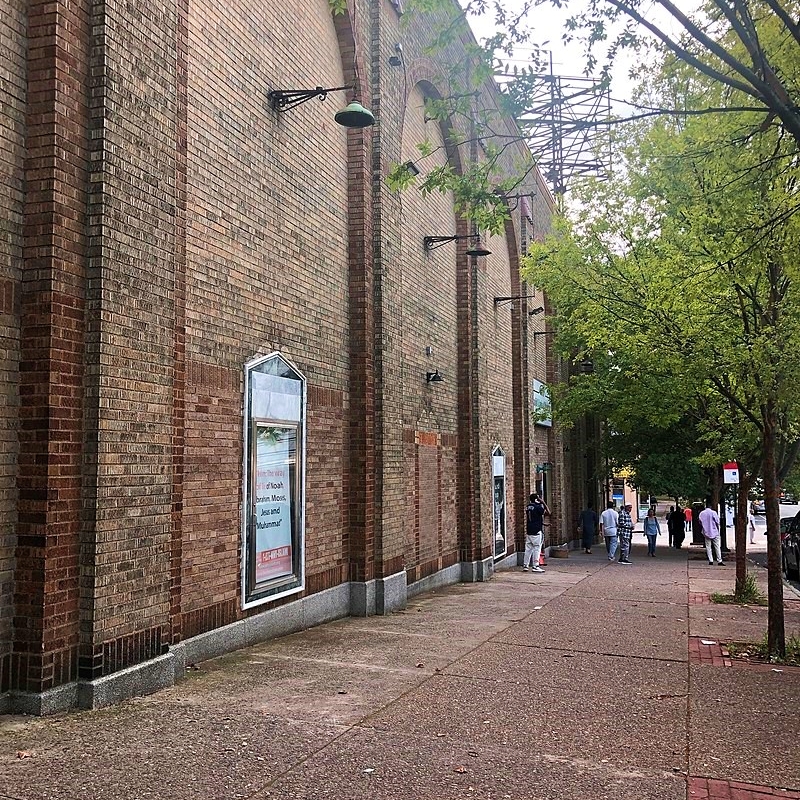 Contemporary color photo of the Masjid Al-Jamia mosque’s street facade. Reddish brown brick with five or six recessed bricked arches shown in the photo. In the foreground is a glass showcase embedded in the wall. Trees line the sidewalk.