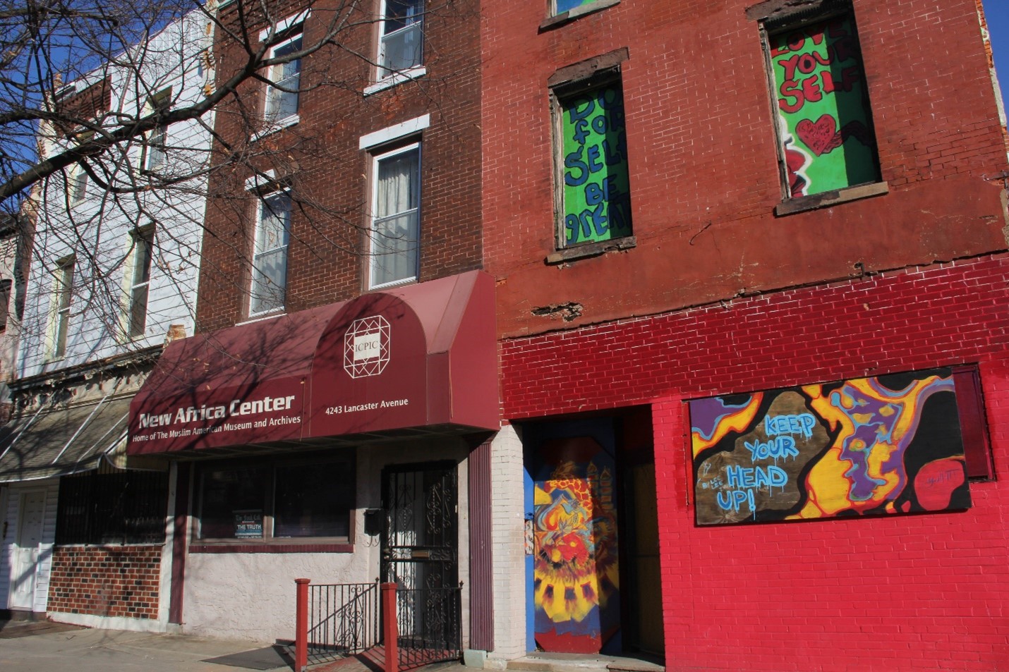 Contemporary color photo highlights the middle building in a row of narrow three-story buildings. This brownish-red brick structure houses the New Africa Center. A red-painted wooden canopy covers the entrance to the first floor. The name NEW AFRICA CENTER is painted in large white letters on the canopy. Smaller case letters some six inches below the name announce the building as home to the Muslim American Museum & Archive.