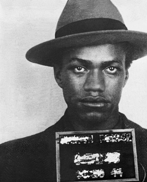 Black and white of late-adolescent Malcolm Little, wearing fedora with a black band, looking directly at the police camera.