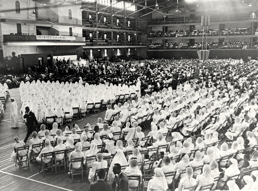 Black and white photo from 1965, a massive indoor venue with thousands of Nation of Islam followers. The foreground to center of the photo shows several hundred women wearing white abayas, seated in folding chairs. A banner hung in the far-left balcony reads THERE IS NO GOD BUT ALLAH AND MUHAMMAD IS HIS PROPHET.