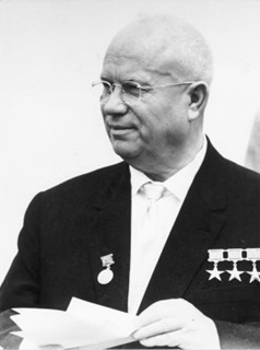 Black and white photo of Nikita Khrushchev in 1963. Khrushchev is distinguished by his bald head and bulbous face. He wears a dark suit and dark tie; a row of medals appears chest-high on the right side of his jacket. He holds assorted papers in his hands.