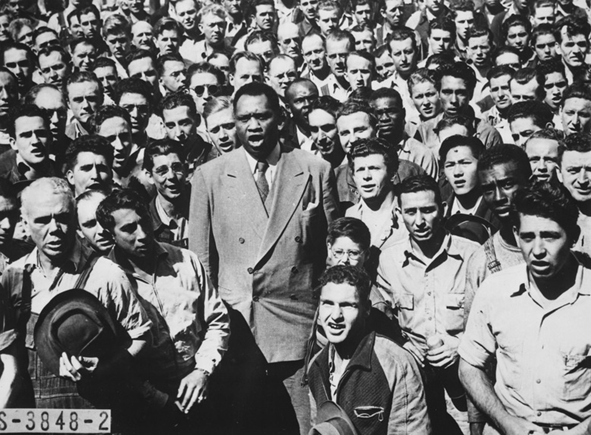 Black and white photo. A singing Paul Robeson stands tall in the foreground of this photo, surrounded by a crowd of all-male shipyard workers. Robeson wears a double-breasted, light-colored wool suit; both he and the shipyard workers are hatless.
