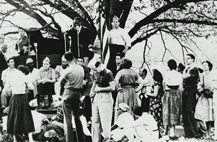 Black and white photo. Paul Robeson is shown preparing to speak at a standing microphone on a stage located in front of a large, multibranched tree. Robeson wears a suit. On and below the stage are five male bodyguards wearing light-colored garrison hats. Hatless women and men comprise the rest of the audience in the photo.