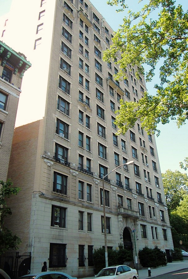 Contemporary color photo of 555 Edgecombe Ave. A multistoried stone building of beige coloring, viewed from the street in late springtime, the season suggested by the bright leafy trees that appear in the image. This otherwise unremarkable building’s only distinctive architectural feature is the Romanesque arch that frames the entrance and includes a small decorative balcony above the arch.