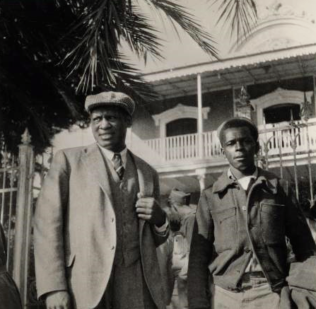 Black and white photo of Paul Robeson, shown in the foreground standing beneath a palm tree beside a young African American man who is a volunteer with the International Brigade in the Spanish Civil War. Robeson wears a light-colored suit with vest, striped tie, and English cap. The volunteer wears a dark, buttoned jacket. In the background is a Spanish colonial-style building with a white-wood balcony railing.