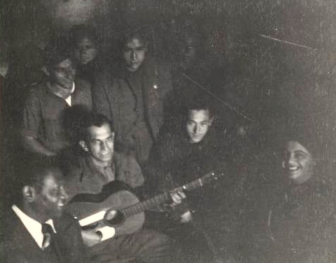 Black and white photo of Paul Robeson singing with freedom fighters at the Madrid battlefront in 1938. Robeson is seated in the lower-left corner of the image beside a man playing a guitar.