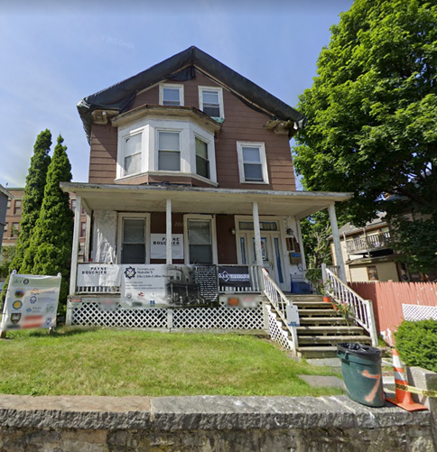 Contemporary color photo of the Roxbury house, showing a three-story working-class domicile with a low stone wall, six stairs with white railings leading up to the front door. The green yard is closely mowed and a large tree in green summer foliage fills much of the right side of the image.