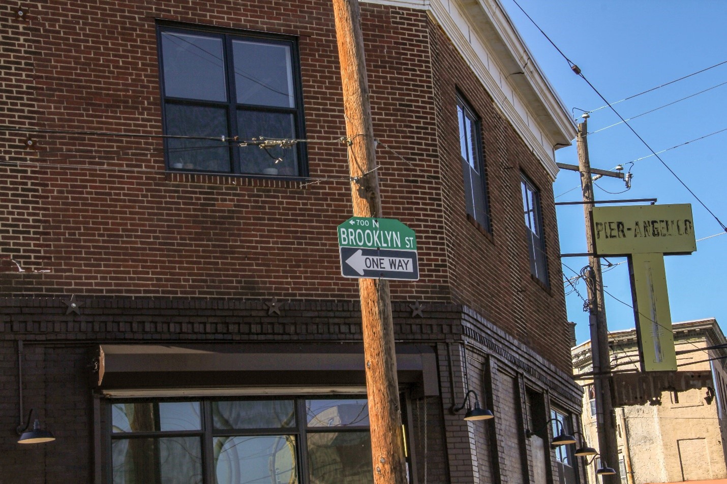 Color photograph of a two-story red brick building at a street corner. A street sign says, “700 N, BROOKLYN ST” and a ONE WAY sign are nailed to a telephone pole.