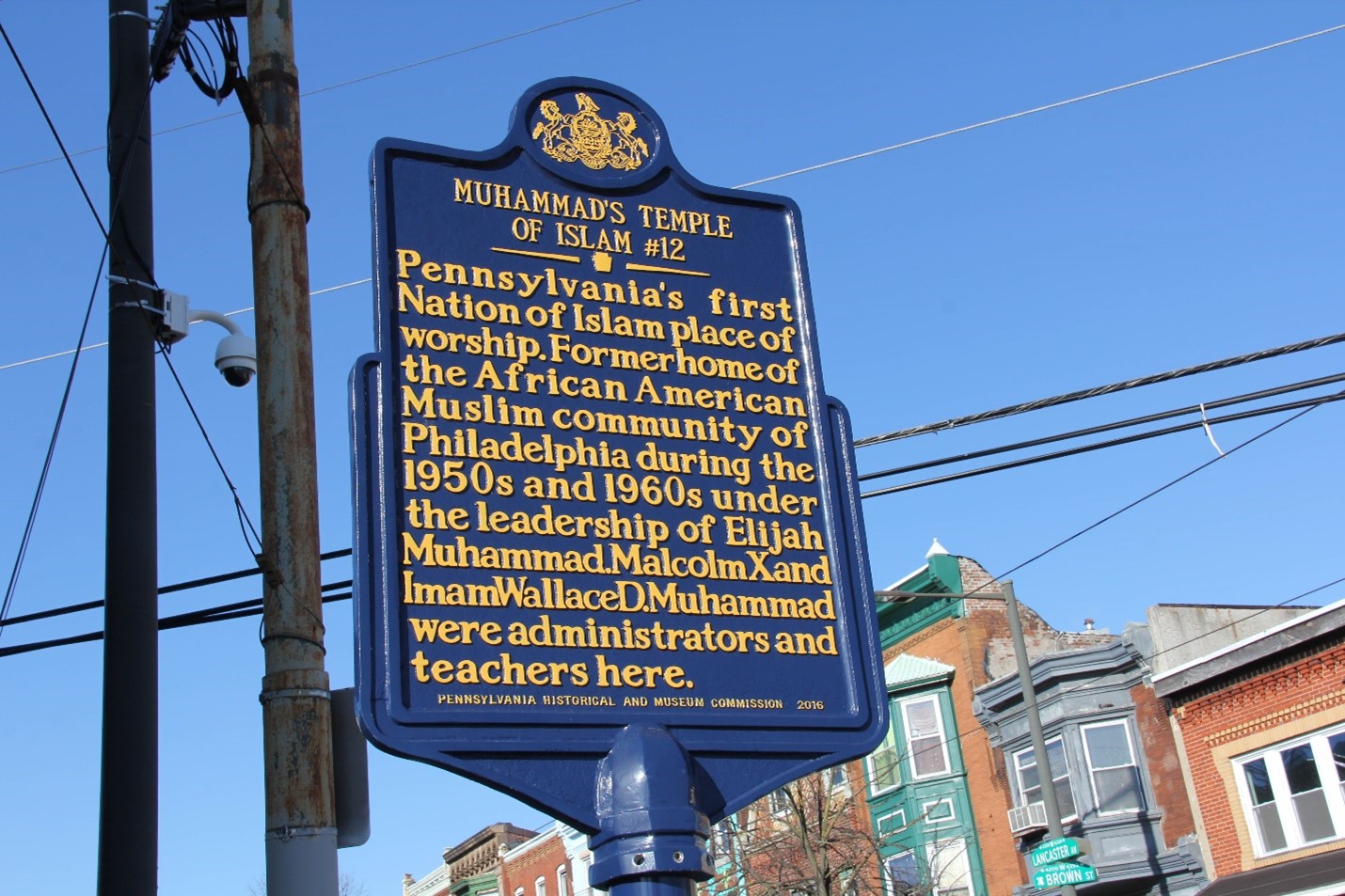 Color photo of a historical marker on a street with modest brick row homes at the intersection of LANCASTER ST and BROWN ST, which reads: MUHAMMAD’S TEMPLE OF ISLAM #12| Pennsylvania’s first Nation of Islam place of worship. Former home of the African American Muslim community of Philadelphia during the 1950s and 1960s under the leadership of Elijah Muhammad. Malcolm X and Imam Wallace D. Muhammad were administrators and teachers here. - Pennsylvania Historical and Museum Commission 2016