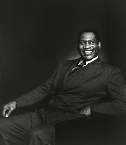 Iconic black and white photo portrait of a smiling Paul Robeson at roughly age 43, seated at an angle facing the camera, with both legs on the floor and his left arm on his chair’s armrest. He wears a dark suit jacket with wide lapels.