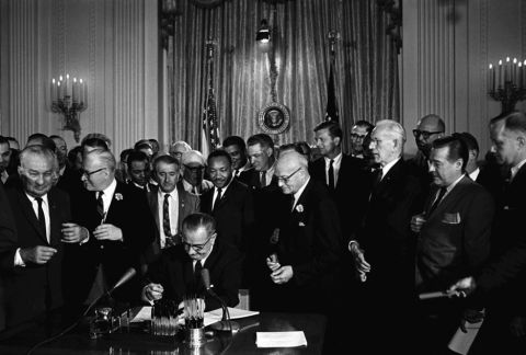 Black and white photo shows a seated President Lyndon B. Johnson surrounded by male dignitaries, most of whom are white, as he signs the 1964 Civil Rights Act. Dr. Martin Luther King Jr., one of the few Black people in the photo, stands directly behind the president to view the signing.