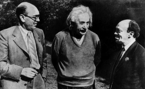 Black and white photo. Balding, bespectacled Izik Feffer stands at left wearing an unbuttoned light gray-tone suit and grey-tone striped tie. Albert Einstein, distinguished by his unruly white hair and the white mustache that covers his upper lip, stands at center wearing a dark gray-tone, v-neck wool sweater. A balding Solomon Mikhoels stands at right, shown wearing a dark suit. The men are smiling and engaged in conversation.