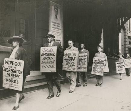 Black and white photo. Paul Robeson shown marching in front of the theater. Like others in the demonstration, he carries a protest sign hung from his shoulders. His sign reads FORDS DISCRIMINATES AGAINST AMERICAN CITIZENS. Robeson appears behind a woman in an oval hat carrying a sign protesting racial segregation at Fords Theater; he wears a dark suit and a fedora.
