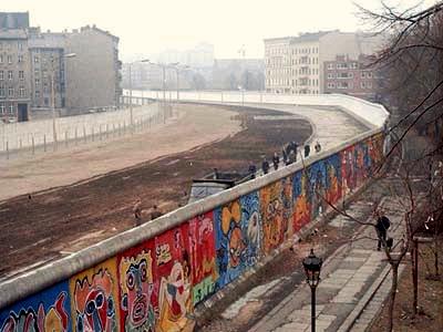 Color photo of a section of the Berlin Wall in the winter of 1986. This image shows the colorful murals of the outer wall, painted by West Germans on their side of the border. The infamous Death Strip appears below and beyond the outer wall. The Death Strip meets an interior wall that provides a barrier to potential fugitives. Bleak, gray, shuttered apartment buildings hover over the interior wall.