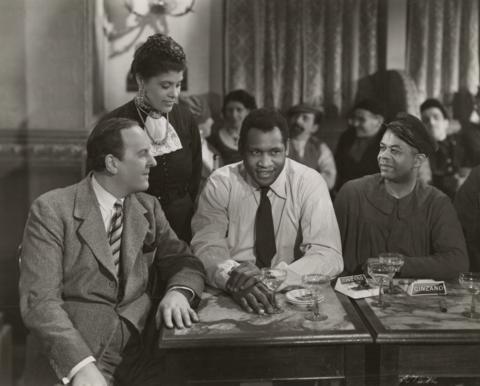 Black and white photo still from the 1938 film Big Fella, showing Paul Robeson seated in a restaurant, with his wife, Eslanda Goode Robeson, standing behind him, and his pianist Lawrence Brown seated on his left. Robeson wears a white shirt with a dark tie.