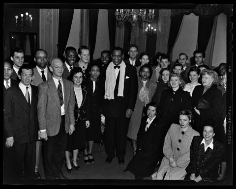 Black and white photo. A smiling Paul Robeson stands beneath a candelabra surrounded by a racially integrated group of men and women at the California Labor School. He wears a black tuxedo with black tie, and a scarf around his neck.