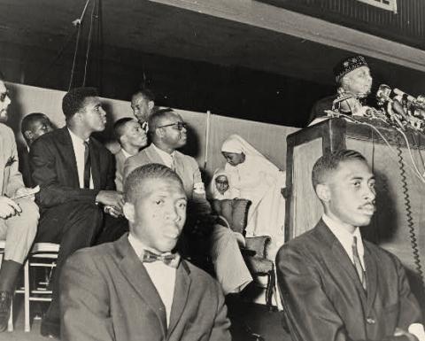Black and white photo. Cassius Clay, soon to adopt the name Muhammad Ali, is seated behind two Fruit of Islam guards. He looks toward the speaker podium where Elijah Muhammad addresses an audience. Elijah Muhammad wears a male Muslim prayer hat. Clay wears a dark suit, white shirt, and dark tie, without a hat.
