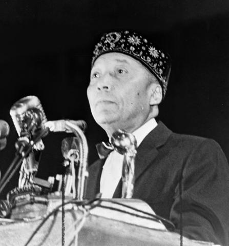 Black and white photo of Elijah Muhammad standing behind a podium with several microphones. He is wearing a white shirt with a bow tie, a dark suit jacket, and an embroidered dark brimless hat. He is cleanshaven.