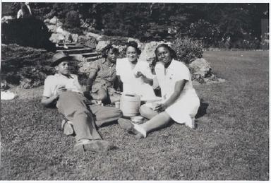 Black and white photo of young Malcolm, clad in summer clothes and a fedora, relaxing in a yard with three women sitting on the ground beside him. Ella Little Collins is on the far right; she wears a short-sleeve white dress.