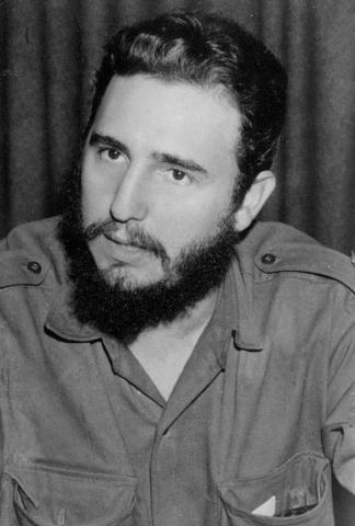 Black and white photo of Fidel Castro with dark hair and beard wearing fatigues.