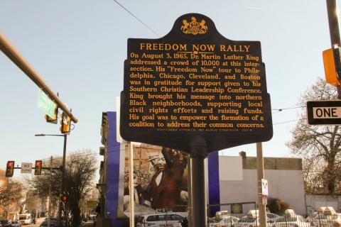 This black, metallic historical marker stands near the intersection of 42nd and Lancaster. It is embossed with gold lettering. The marker’s heading, FREEDOM NOW RALLY, appears in a larger print-font than the eleven lines of brief explanatory content below the heading.