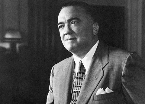 Alt text: Black and white photo of J. Edgar Hoover wearing a patterned tie, white shirt, mid-tone suit jacket with a white pocket handkerchief