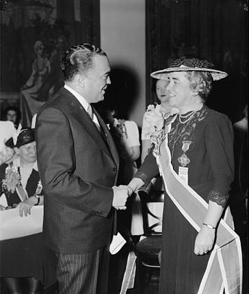 Black and white photo of J. Edgar Hoover shaking hands with a middle-aged woman. Hoover is dressed in a dark suit, and the woman is wearing a straight dark dress, a large oval hat, a pearl necklace, and a large sash that flows from her right shoulder to a point on her torso below her left hand.