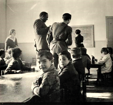 Black and white photo of Paul Robeson and his Spanish guide, Captain Castillo, at the Karl Marx School in Madrid, in 1938, with Robeson and Castillo in the middle ground and two young boys seated at a table, facing the camera, in the foreground.