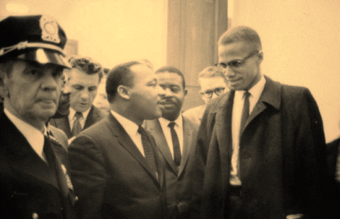 Sepia-tone photo showing Dr. Martin Luther King Jr. and Malcom X in conversation in the U.S. Capitol building. Dr. King wears a dark suit, white shirt, and dark tie; Malcolm X wears an open overcoat with a dark tie. At left in the image stands a white Capitol policeman. Rev. Ralph Abernethy stands behind Dr. King.