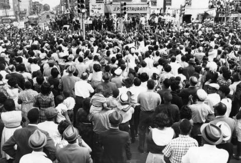 Black and white photo showing a large crowd of men, women, and children in the intersection at 40th and Market St., congregating for a rally led by Dr. Martin Luther King Jr.  Dr. King and other Black leaders stand on a platform in front of the JP RESTAURANT, on the north side of the intersection.