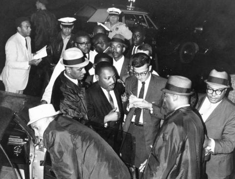 Black and white photo showing Dr. Martin Luther King Jr. surrounded by a group of Black male colleagues on his arrival at Philadelphia International Airport. Several of the men wear fedoras. Dr. King wears a dark suit and light gray-tone tie. He is talking with a white reporter, who is taking notes. Dr. King’s close associate Andrew Young is attired in a light suit and dark tie and appears in the left background.