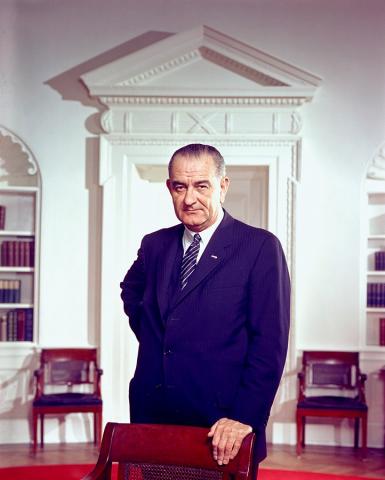 Color photo of President Lyndon Baines Johnson, shown facing the camera and standing with his left arm resting on a chair. He wears a dark blue suit, white shirt, and blue tie with pink stripes. The room is apparently a library of sorts. It is painted white with a Greco-Roman decorative design.