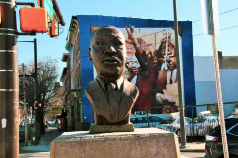 Contemporary color photo of the bronze bust of Dr. Martin Luther King Jr. at 42nd and Lancaster. This figurative sculpture of Dr. King’s head and shoulders, mounted on a stone pedestal, appears in the foreground. A colorful mural of Dr. King with his arms raised at the “Freedom Now Rally” rises above a small parking lot in the background.