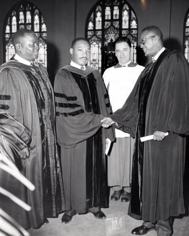Black and white photo of Dr. Martin Luther King Jr. standing between two Black pastors. Dr. King and Rev. Shepard Sr. wear dark academic gowns. Rev. Shepard Jr. wears a traditional black ministerial robe; he and Dr. King shake hands. A fourth person is a white priest, robed in a white gown with a dark clerical collar. Stain-glass windows form the backdrop of the photo.