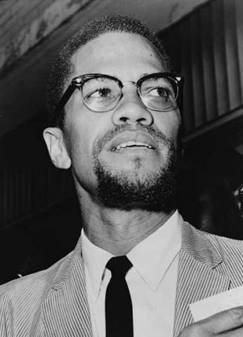 Black and white photo of Malcolm X in 1964. He is standing, wearing a light corduroy jacket, a white shirt, and a dark tie. His head is tilted to his left side. Emblematic of his break with the Nation of Islam, he now has a trimmed moustache connected to a small goatee, forming a circle.