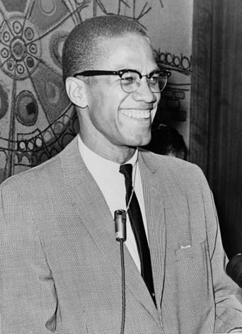 Black and white photo of Malcolm X in 1964. Shown smiling and relaxed, he stands at a microphone. He wears a light gray-tone suit or sports coat, with a dark tie. He has closely cropped hair, is clean shaven, and wears glasses.
