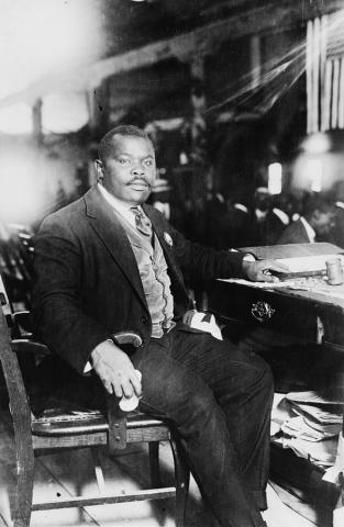 alt text=Black and white photo of Marcus Garvey. He is seated and his chair is turned away from the desk to face the camera. He is dressed in a dark suit, with a buttoned light gray-tone vest and striped tie.
