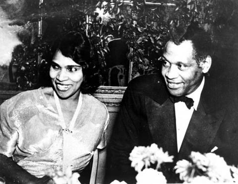 Black and white photo of Paul Robeson and Marian Anderson, seated at a table decorated with a flower arrangement. Robeson, at right in the photo, wears a dark tuxedo with black tie; Anderson’s torso is covered by a formal white blouse. Both Robeson and Anderson are smiling.